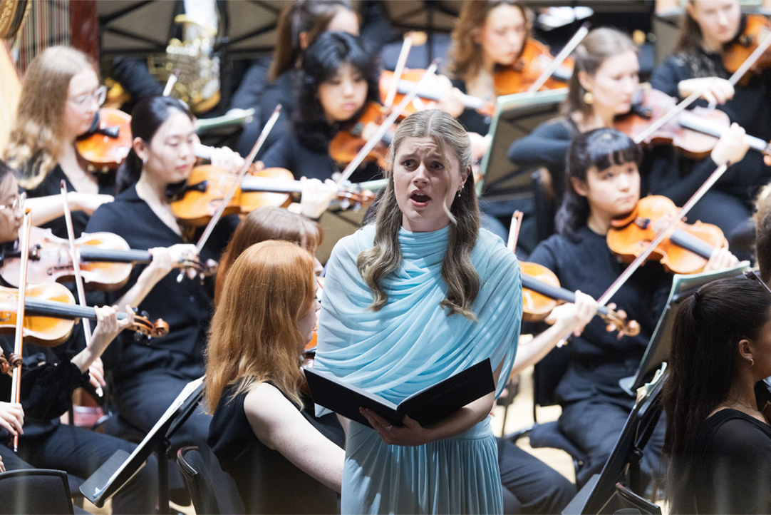 A women wearing a blue dress, singing with a song book open, with an orchestra behind her.